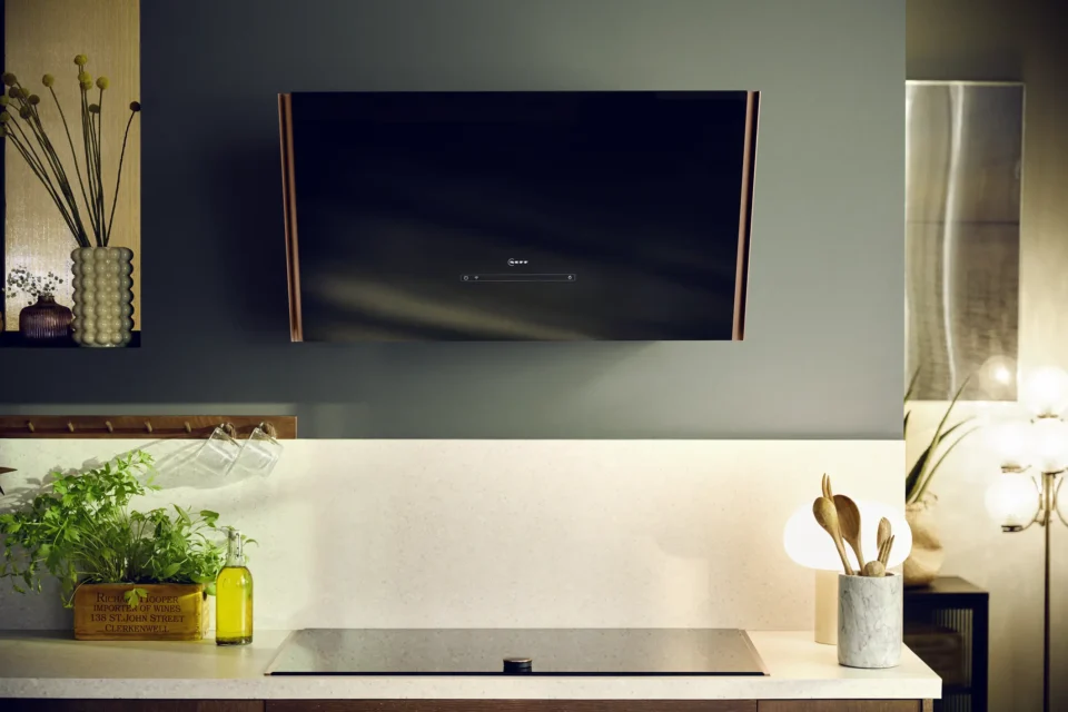 Eine moderne Küche mit einer schwarzen Dunstabzugshaube an der Wand über einem Kochfeld. Auf der Arbeitsfläche stehen Kräuter in einer Holzkiste, daneben Küchenutensilien in einem Behälter und eine Lampe mit rundem Schirm.
