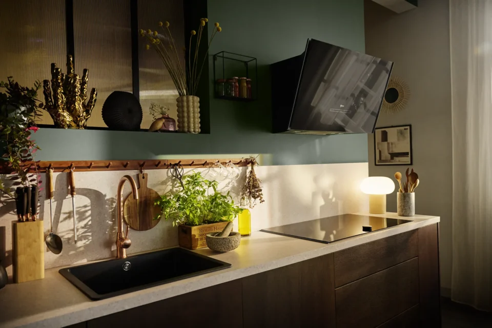 Moderne Küche mit dunklen Holzschränken. Eine Arbeitsplatte mit Spüle, Kräutertopf, gelber Flasche und Beleuchtung. Links Messerblock, Trockenblumen und Gitterregale an grüner Wand. Rechts Kochfeld und helle Lampe.