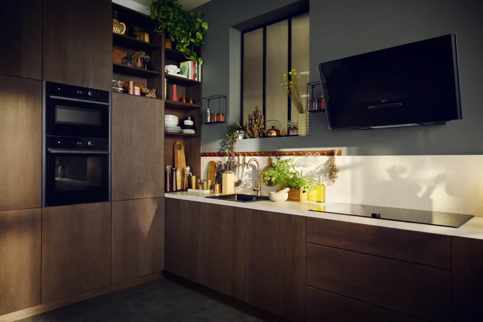 Moderne Küche mit dunklen Holzschränken, eingebauten Geräten und dünnen Regalen. Eine Arbeitsplatte mit Spüle und Kochfeld. Pflanzen und Dekorationsgegenstände ergänzen das Ambiente. Eine Wand ist mit einem großen Fenster ausgestattet.