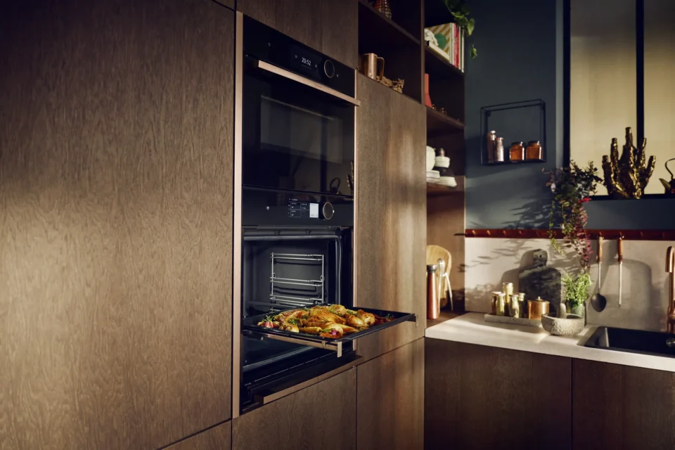 Ein moderner Einbaubackofen in einer Holzküche zeigt eine geöffnete Tür mit einem Backblech voller gebackener Speisen. Die Küche hat Regale mit Pflanzen und Küchenutensilien sowie eine Spüle mit goldenen Armaturen.
