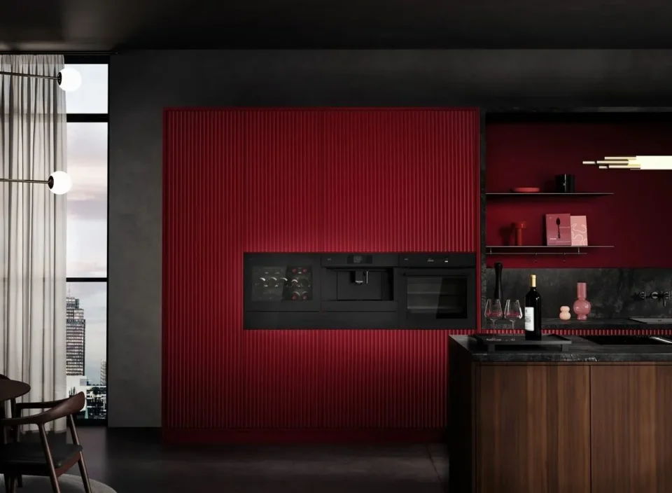 Moderne Küche mit rot geriffelter Wand, eingebauten schwarzen Küchengeräten und einem dunklen Marmortresen mit Weingläsern und Flaschen. Links sind Fenster mit durchsichtigen Vorhängen und städtische Gebäude sichtbar.