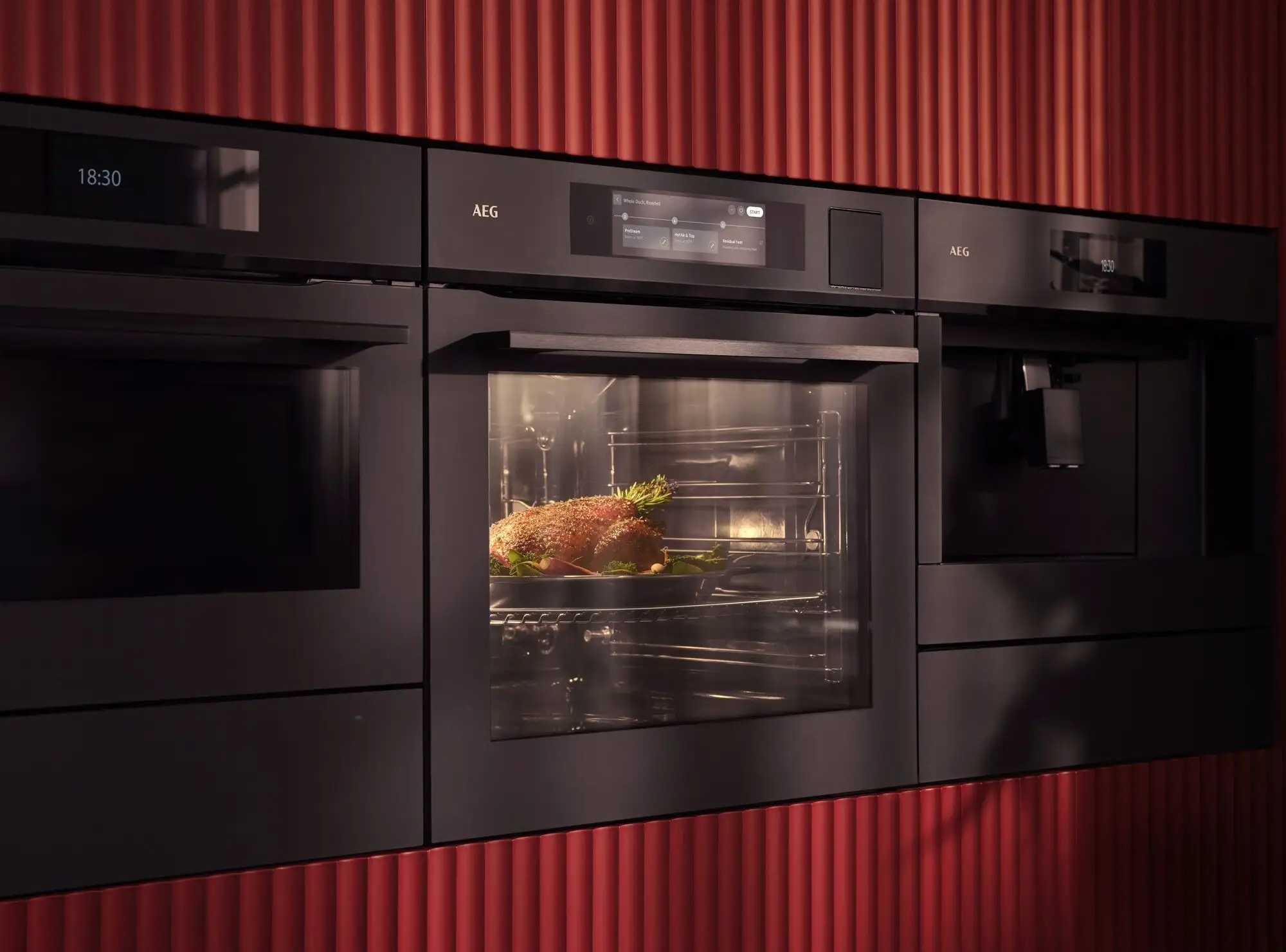 Eine moderne, schwarze Küchengerätewand mit Backofen, Kaffeemaschine und Mikrowelle vor einer gerippten, roten Wand. Im Ofen wird ein Braten mit Rosmarin auf einem Teller gegart.