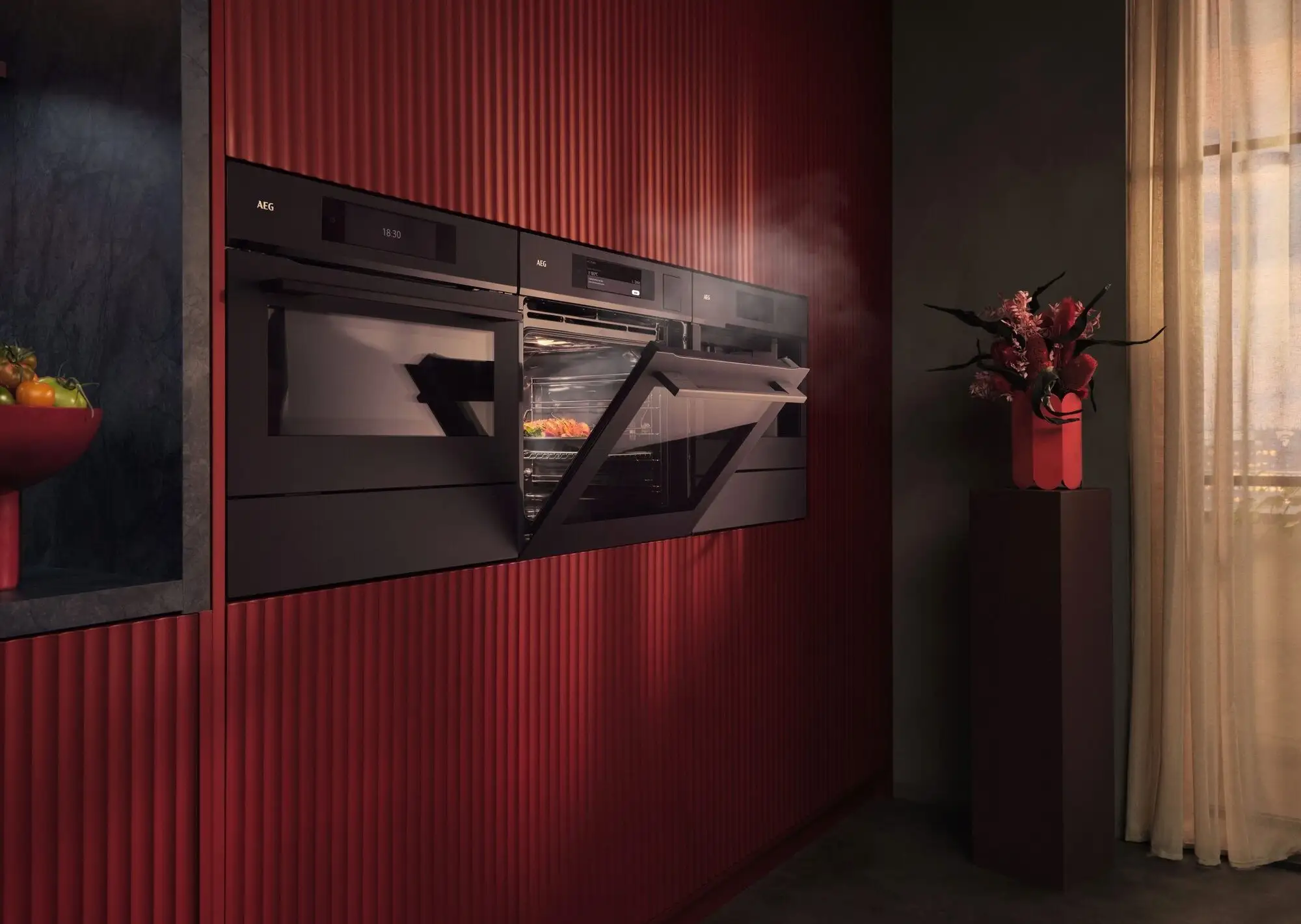Eine moderne, schlichte Küche mit dunkelroten Wänden. Ein Ofen mit leicht geöffneter Tür gibt Dampf ab. Rechts steht eine hohe Vase mit Blumen auf einem Sockel vor einem Fenster mit hellem Vorhang.