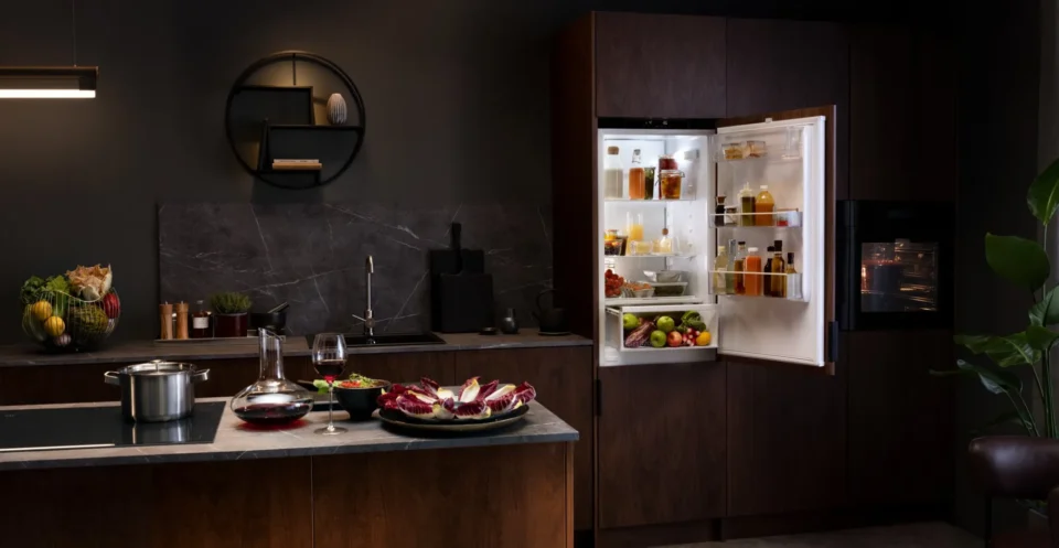 Ein moderner Küchenraum mit einem offenen Kühlschrank, gefüllt mit Lebensmitteln und Getränken. Auf der Kücheninsel stehen eine Schüssel mit Salat, ein Kochtopf und ein Glas Rotwein. Im Hintergrund sind dunkle Schränke und ein rundes Regal zu sehen.