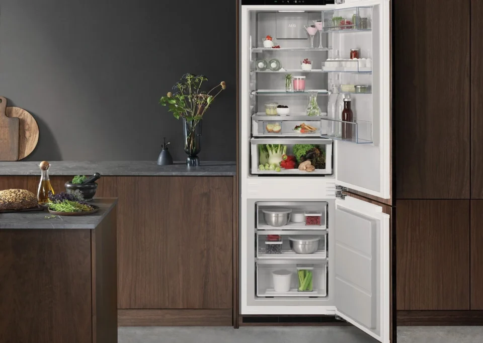 Kühlschrank in einer modernen Küche, geöffnet und gefüllt mit verschiedenen Lebensmitteln wie Gemüse, Joghurt, Saucen und Getränken. Die Küche hat dunkle Holzschränke und eine Arbeitsplatte mit Dekoration.