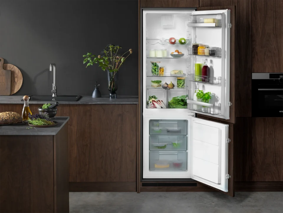 Ein geöffneter Kühlschrank in einer modernen Küche ist gefüllt mit verschiedenen Gemüsen, Getränken und Lebensmitteln. Die Küche hat dunkle Holzschränke, eine Spüle mit Wasserhahn und eine Topfpflanze daneben.