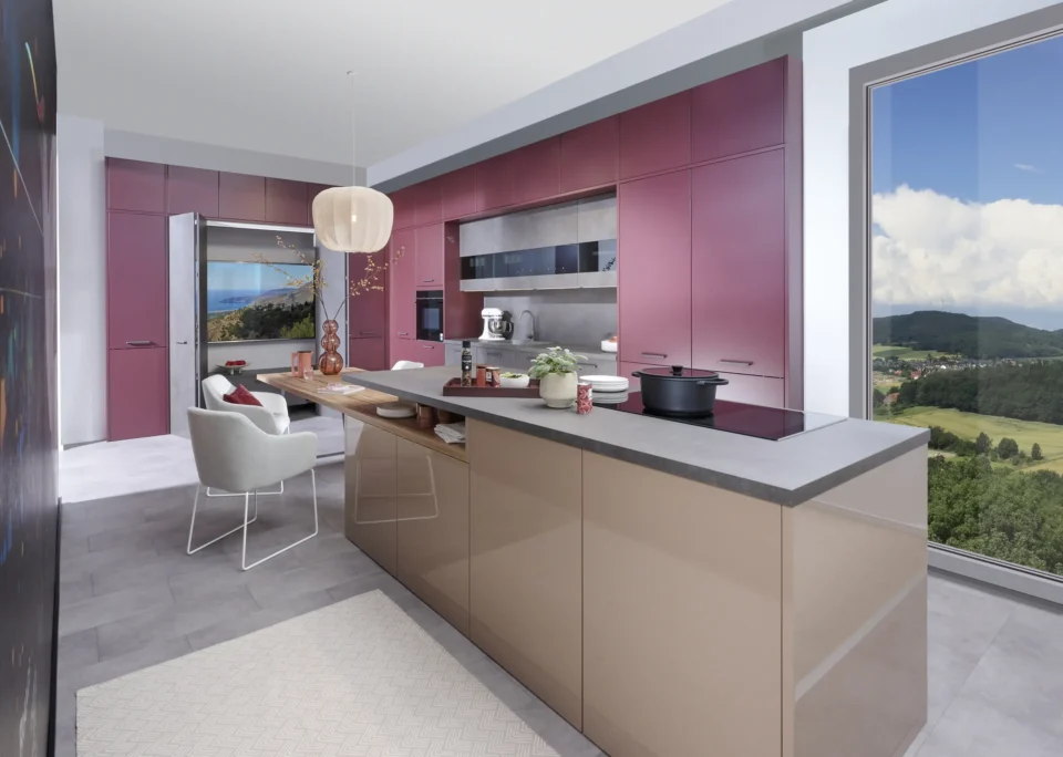 Moderne Küche mit rosa und beige Schränken, grauer Arbeitsplatte, eingebautem Ofen und Herd. In der Mitte steht ein Esstisch mit Stühlen. Das Fenster zeigt eine Landschaft mit Hügeln und einem bewölkten Himmel.
