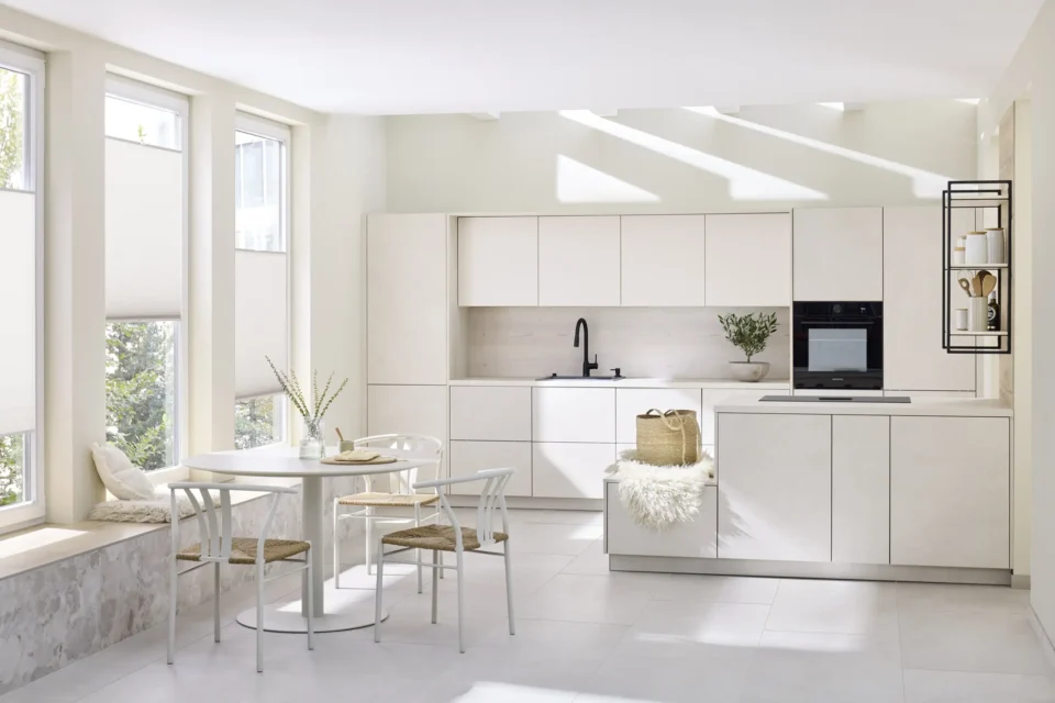 Helle Küche mit weißen Schränken und Arbeitsplatten. Links Esstisch mit vier Stühlen, auf Fensterbank Dekoration. Rechts eingebaute Geräte. Heller Boden, große Fenster mit Lichtstrahlen. Skandinavisches Design.