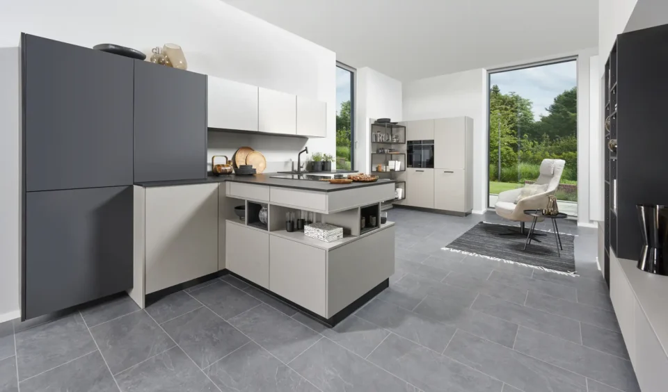 Moderne Küche mit grauen Schränken, einer Insel und integriertem Backofen. Große Fenster bieten Blick auf einen Garten. Eine gemütliche Sitzecke mit Stuhl und Teppich befindet sich im Hintergrund.