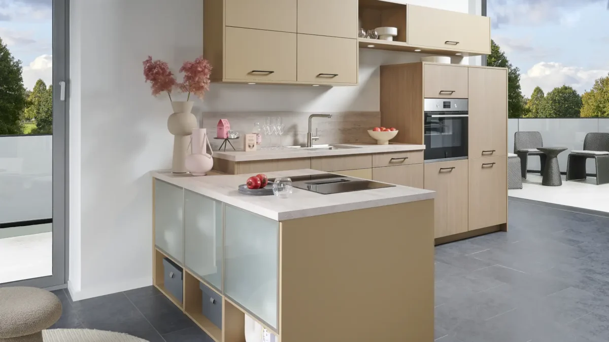2229 2336 A10 Moderne Küche mit L-förmiger Arbeitsplatte, hellbraunen Schränken und Glasfronten. Dekoration mit rosa Blumen und Vasen. Rechts integrierter Backofen, Fenster mit Blick auf Garten. Fußboden aus grauen Fliesen, runder Teppich und Sitzhocker.