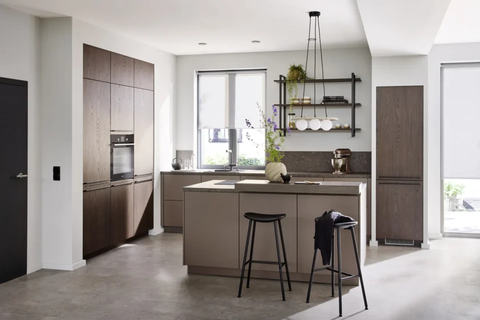 Moderne Küche mit Insel, dunklen Holzschränken, eingebautem Ofen und Hänge-Glaslampen. Zwei hohe Fenster lassen Tageslicht herein. Ein schwarzer Barhocker steht an der Insel, Dekorationen auf der Arbeitsfläche und Regalen.