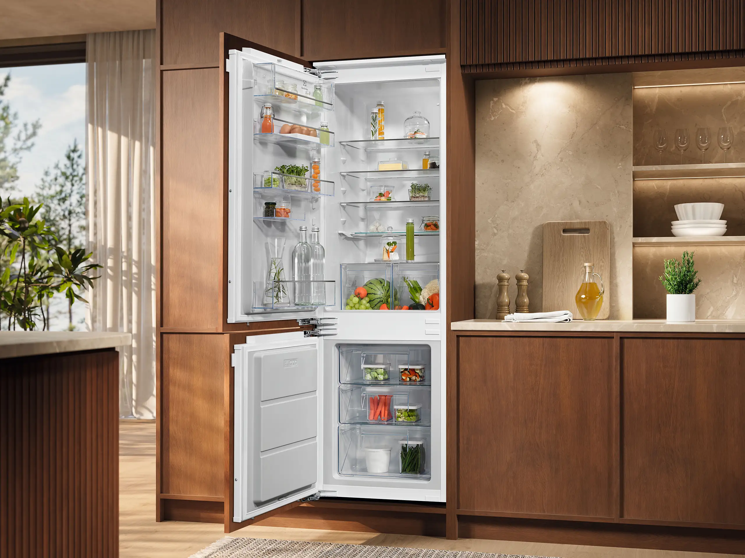 Ein offener, moderner Kühlschrank in einer stilvollen Küche. Der Kühlschrank ist gut organisiert mit Gemüse, Getränken und anderen Lebensmitteln. Daneben steht ein Schneidebrett mit einer Flasche Öl und Dekoration.