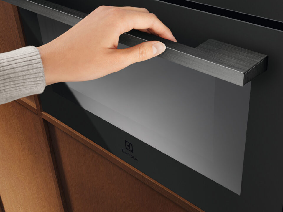 Eine Hand öffnet die Tür eines modernen Backofens mit Edelstahlgriff. Der Backofen ist in einen Holzschrank eingebaut und die Person trägt einen hellgrauen Pullover.