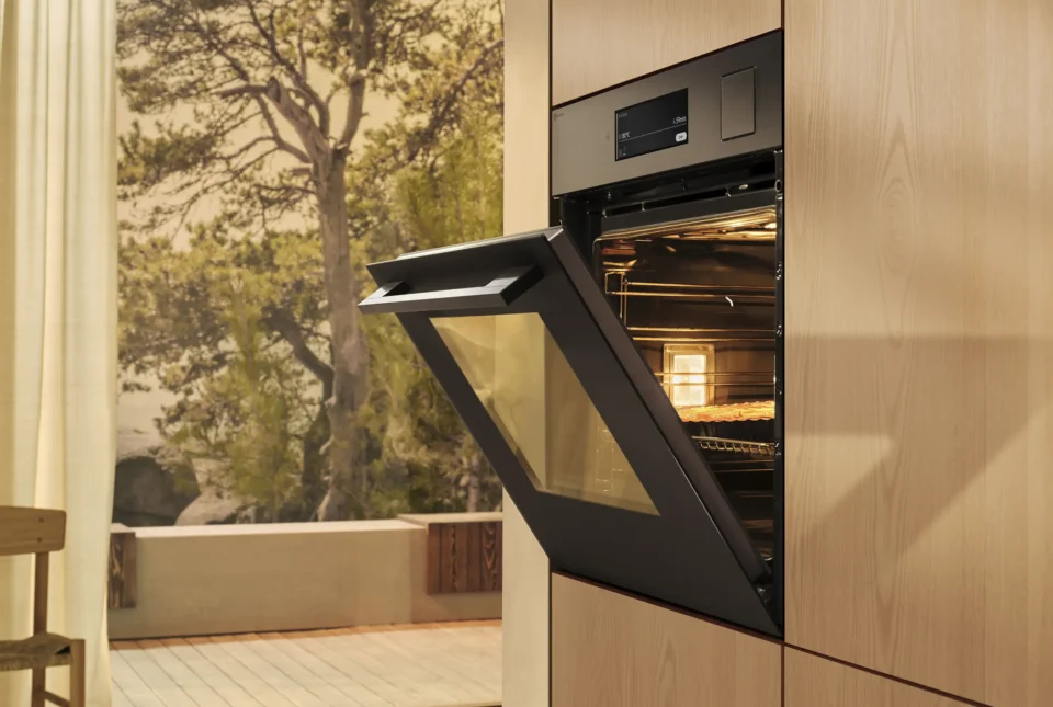 Ein moderner Backofen ist in eine holzverkleidete Wand integriert. Die Ofentür steht offen, und innen befindet sich ein Backblech mit Backgut. Im Hintergrund ist eine Wand mit Naturmotiv zu sehen.
