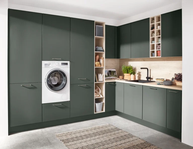Moderne Küche mit grünen Schränken, einer eingebauten Waschmaschine, offenem Regal mit Deko, Arbeitsplatte aus Holzoptik, schwarzer Armatur und Spüle. Grauer Bodenbelag und rechteckiger Teppich im Vordergrund.