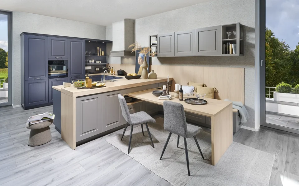 Moderne Küche mit grauen Schränken und einem Holz-Esstisch, der in zwei gepolsterte Stühle übergeht. Dekorationen und Pflanzen verteilen sich auf den Oberflächen, große Fenster bieten Blick ins Grüne.