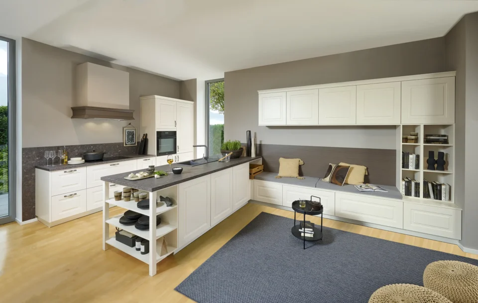 Helle, moderne Küche mit L-förmiger Arbeitsplatte, weißer Schränken, grauem Teppich und Holzfußboden. Links ein Fenster, Sitzbank mit Kissen und Regalen rechts. Küchenutensilien sind auf der Arbeitsplatte verteilt.