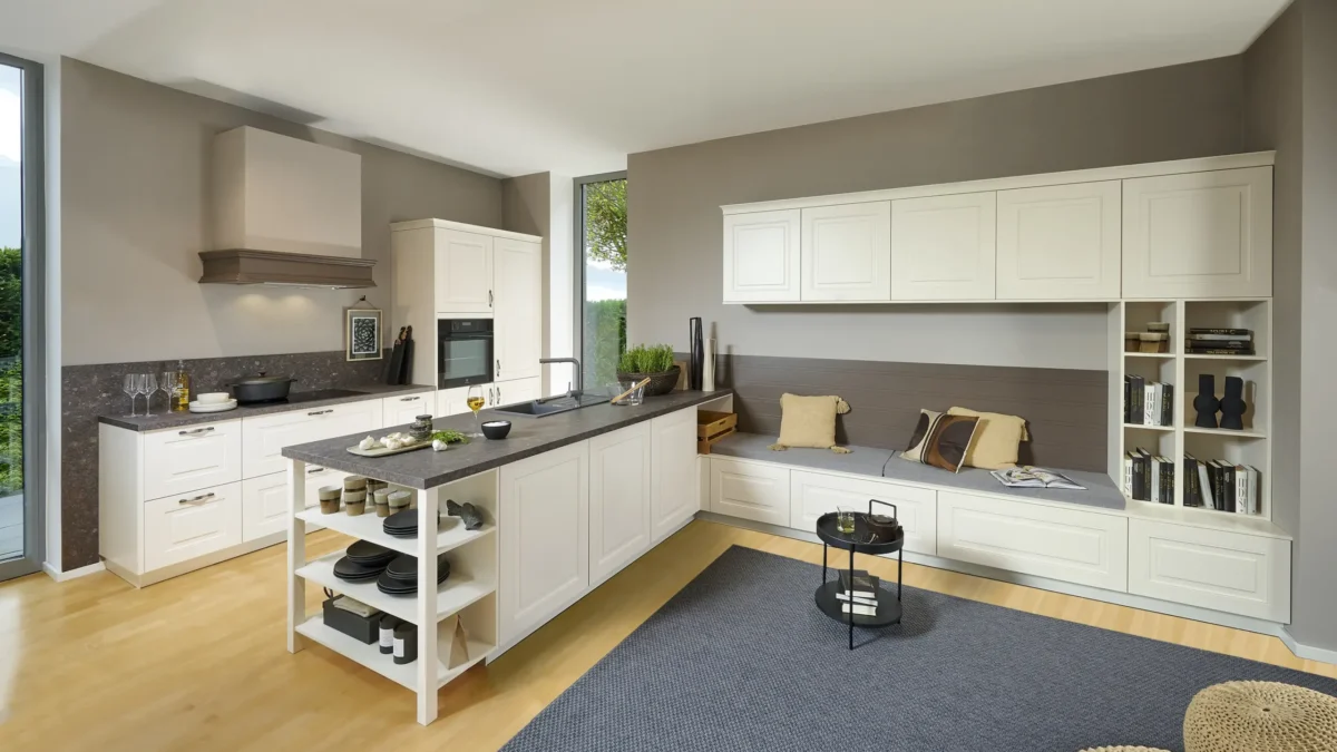 3470 79 A10 Moderne Küche mit Küchenschränken in Cremefarbe, einer grauen Arbeitsplatte und einem integriertem Sitzbereich. Auf der Arbeitsplatte stehen Geschirr und Dekoration. Der Raum wirkt hell und geordnet. Ein großes Fenster lässt Tageslicht herein.