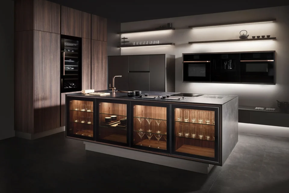 Moderne Küche mit dunkler Holzoptik: Kochinsel mit Glasvitrinen voller Gläser im Vordergrund, eingebauter Backofen und Regale an der Wand im Hintergrund. Gedämpfte Beleuchtung schafft eine elegante Atmosphäre.