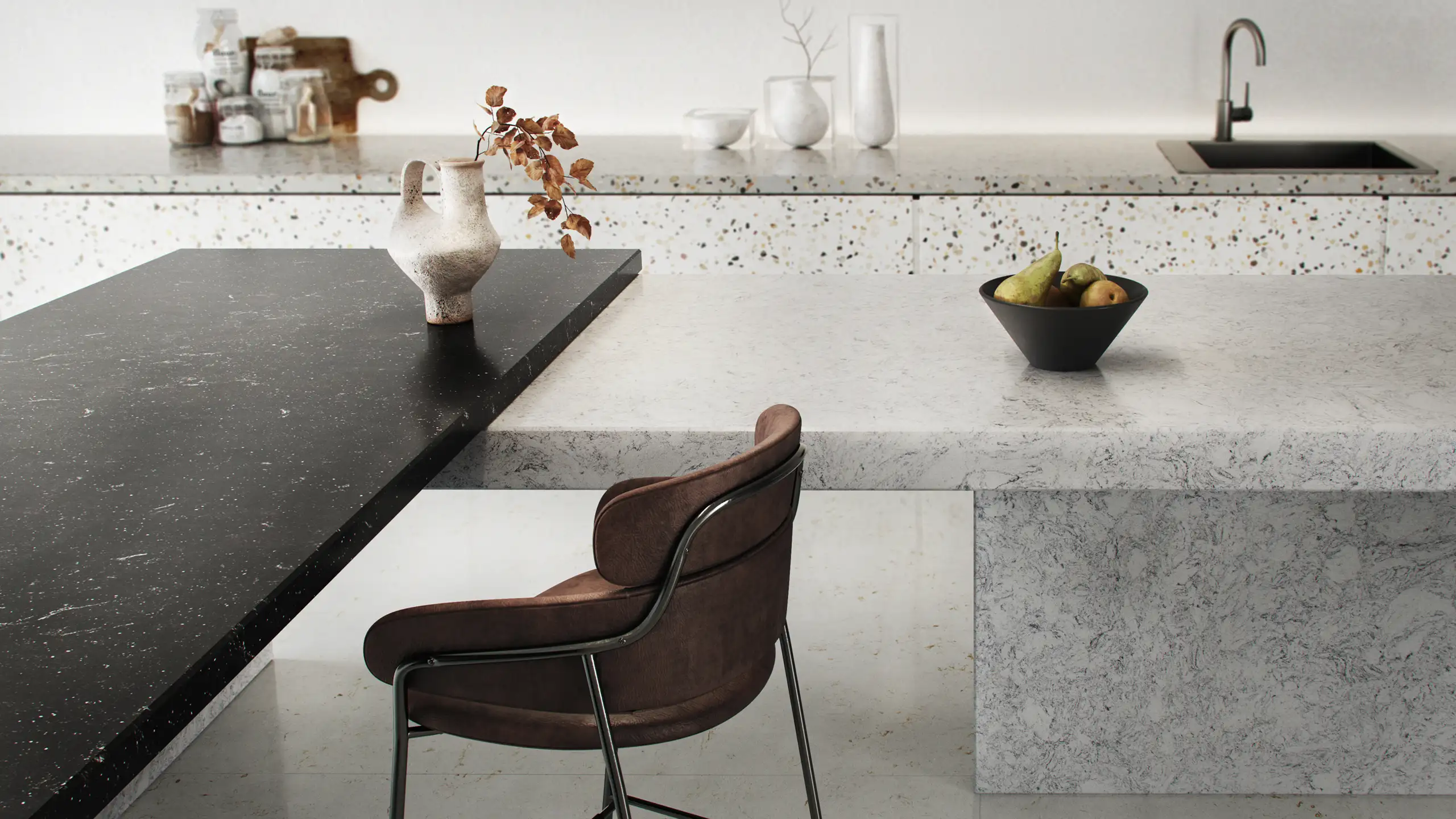 Moderne Küche mit einer schwarzen und einer weißen Arbeitsplatte. Darauf stehen eine Vase mit Zweigen und eine Schale mit Obst. Ein brauner Stuhl ergänzt das minimalistische Design. Im Hintergrund sind Gläser und ein Wasserhahn zu sehen.
