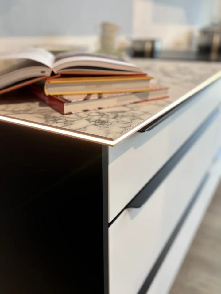 Ein Stapel aufgeschlagener Bücher liegt auf einer modernen Küchenarbeitsplatte mit Marmoroptik. Die Arbeitsplatte hat eine beleuchtete Kante. Darunter befinden sich schwarze und weiße Schubladen.