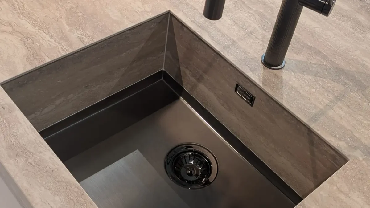 Ein quadratisches Waschbecken aus dunklem Material ist in eine Arbeitsplatte mit heller Steinoptik eingelassen. Darüber befindet sich ein moderner, schwarzer Wasserhahn.