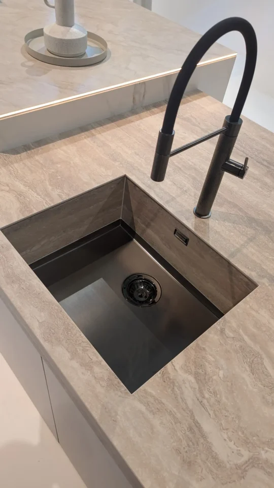 Ein quadratisches Waschbecken aus dunklem Material ist in eine Arbeitsplatte mit heller Steinoptik eingelassen. Darüber befindet sich ein moderner, schwarzer Wasserhahn.