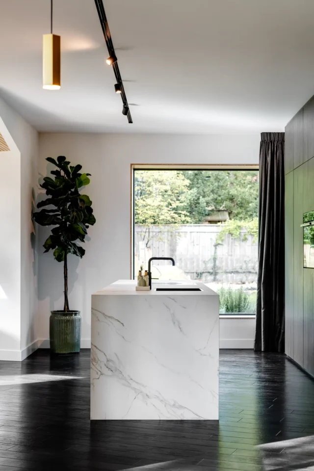 Helle, moderne Küche mit einer weißen Insel aus Marmor. Eine große Fensterfront bietet Blick ins Grüne. Links steht eine große Pflanze in einem Topf. Die Decke hat eingelassene Beleuchtung.