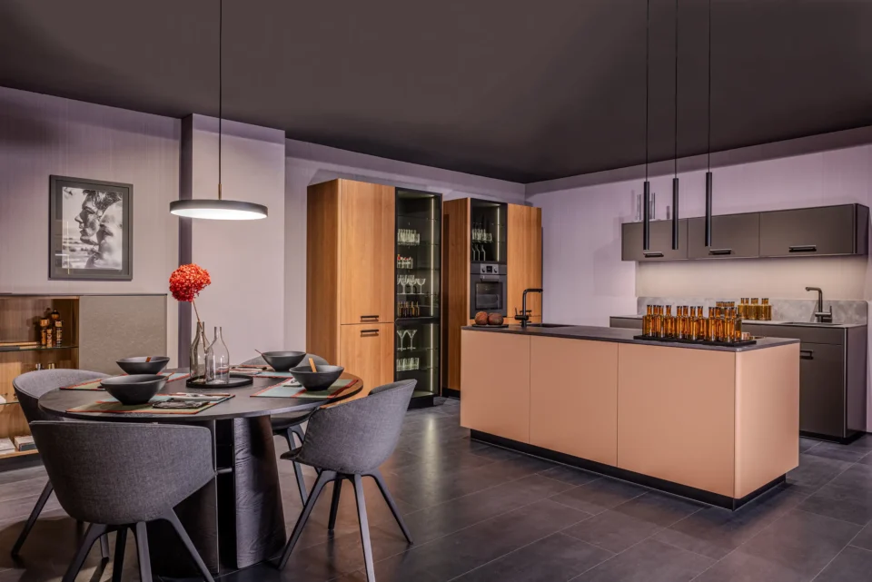 Moderne Küche mit dunklen Fliesen, heller Insel mit Hockern, eingebautem Kühlschrank und Schränken. Runder Esstisch mit grauen Stühlen und roten Blumen. Gedämpfte Beleuchtung von Deckenlampen. Wand mit Kunstwerk.