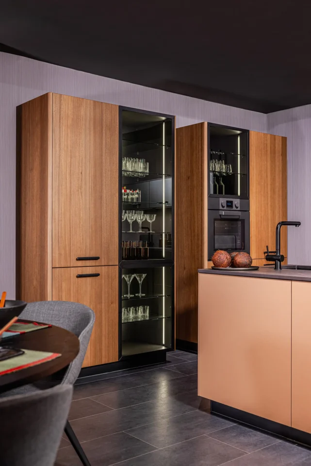 Moderne Küche mit Holzelementen: Zwei hohe Schränke mit Glastüren und beleuchteten Böden zeigen Gläser. Eine Insel mit dunklem Wasserhahn und zwei runden Dekorationen im Vordergrund. Grauer Boden und heller Hintergrund.