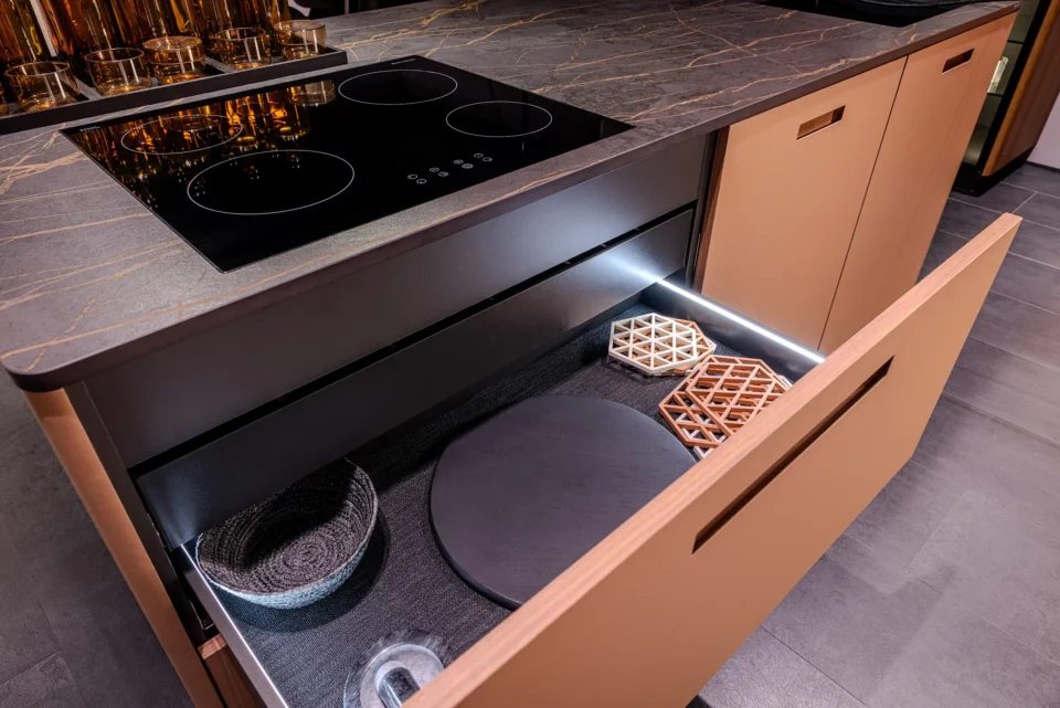 Eine moderne Küche mit einem dunklen, gemusterten Arbeitsplattenherd. Darunter befindet sich eine offene Schublade mit Tabletts und Körben, beleuchtet durch LED-Licht. Der Boden ist grau.