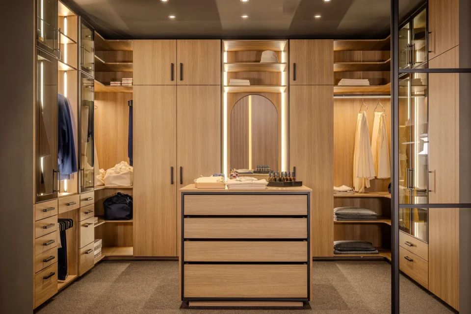 Ein moderner begehbarer Kleiderschrank mit Holzschränken, Schubladen in der Mitte und eingebauter Beleuchtung. Kleidung und Accessoires sind ordentlich aufbewahrt. Ein Spiegel befindet sich an der Rückwand.