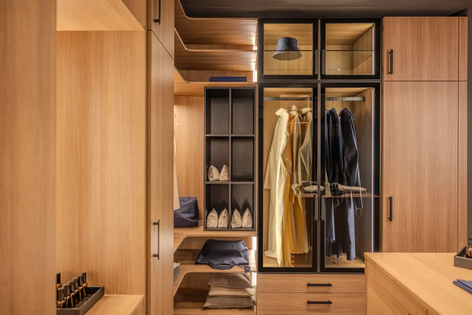 Geräumiger begehbarer Kleiderschrank mit Holzschränken, offenen Regalen und Glastüren. Darin hängen farbige Kleidungsstücke und es sind Schuhe und gefaltete Kleidungsstücke sichtbar. Deckenbeleuchtung erhellt den Raum.