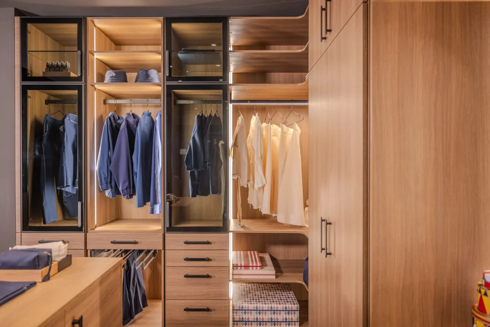 Ein offener, moderner Kleiderschrank aus Holz mit Glasfronten zeigt ordentlich aufgehängte blaue und weiße Kleidungsstücke. Darunter befinden sich Schubladen und gefaltete Textilien. Oben sind Accessoires platziert.