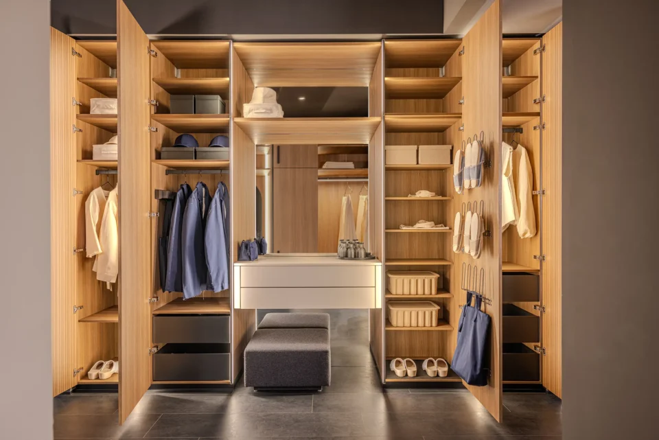 Ein offener Kleiderschrank mit Holzoberfläche, gefüllt mit ordentlich aufgehängter Kleidung, Regalen für Accessoires, Schubladen und einem zentralen Spiegel über zwei weißen Schubladen. Ein grauer Hocker steht davor.