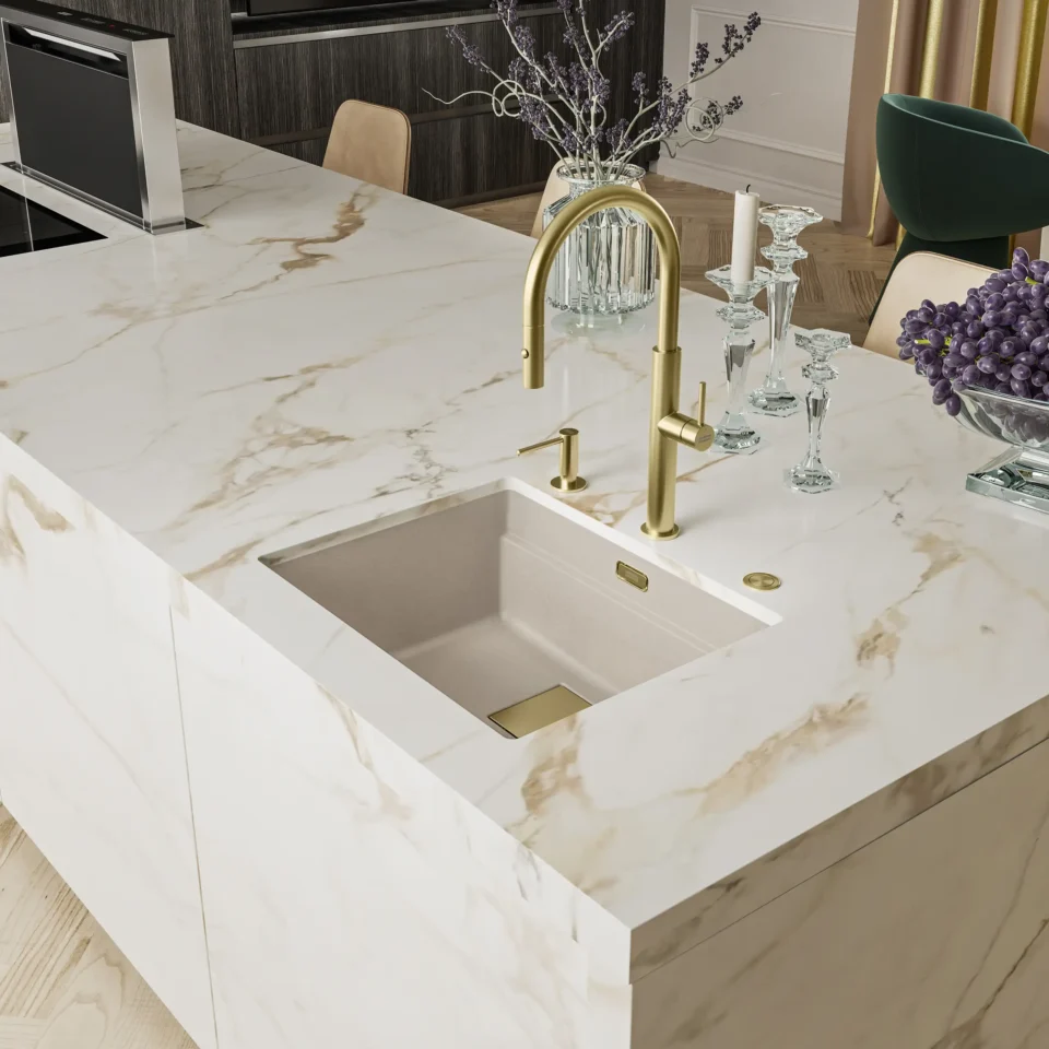 Eine moderne Kücheninsel mit einer weiß-goldenen Marmoroberfläche. Integriert ist ein rechteckiges Waschbecken mit goldenem Wasserhahn. Daneben stehen Kerzenhalter, Weingläser und ein Strauß violetter Blumen.
