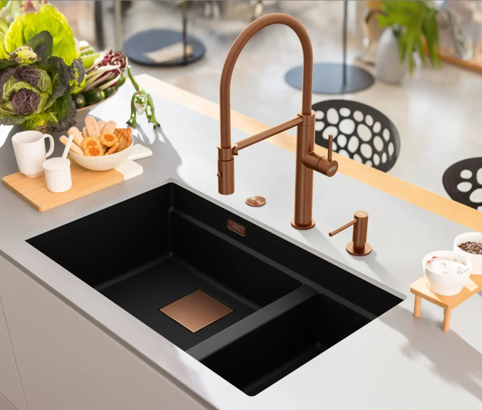 Eine moderne Küche mit einem großen schwarzen Spülbecken und einem kupferfarbenen Wasserhahn. Auf der Arbeitsplatte liegen Obst und Backwaren. Dahinter stehen Stühle mit runden Aussparungen an der Rückenlehne.