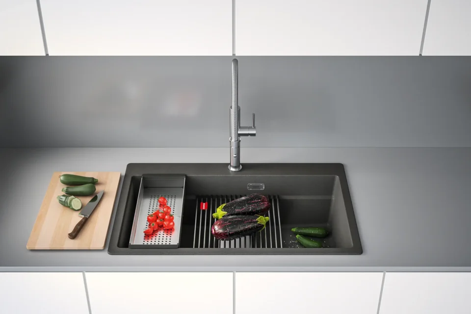 Eine moderne Küchenspüle mit grauer Arbeitsplatte. Links ein Schneidebrett mit geschnittenen Gurken, in der Spüle ein Sieb mit Tomaten und zwei lilafarbene Maiskolben auf einem Gitter. Rechts zwei ganze Gurken.