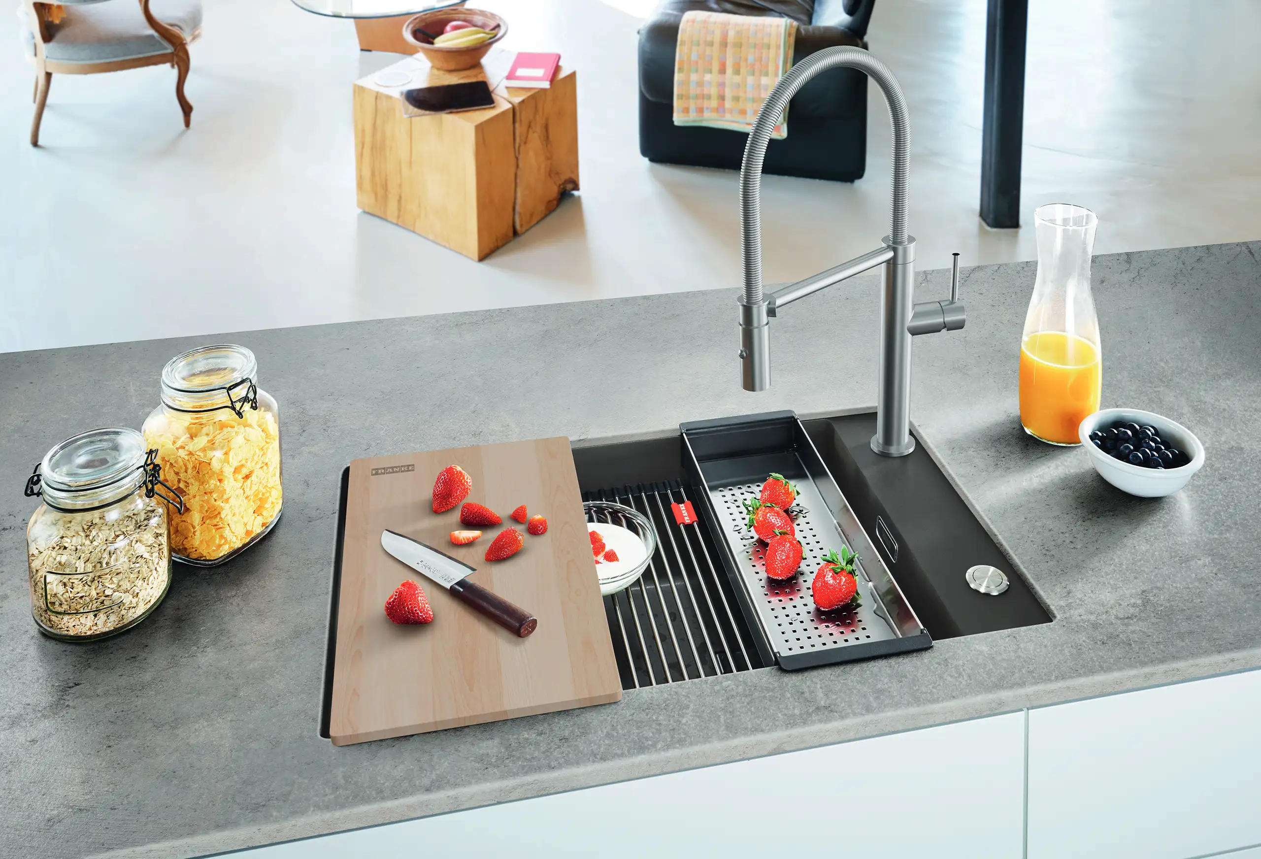 Eine moderne Küchentheke mit einer Edelstahlspüle. Auf der Arbeitsplatte liegen ein Schneidebrett mit Erdbeeren und ein Messer. Daneben sind ein Sieb, ein Glas Saft und zwei Vorratsgläser mit Müsli und Haferflocken.