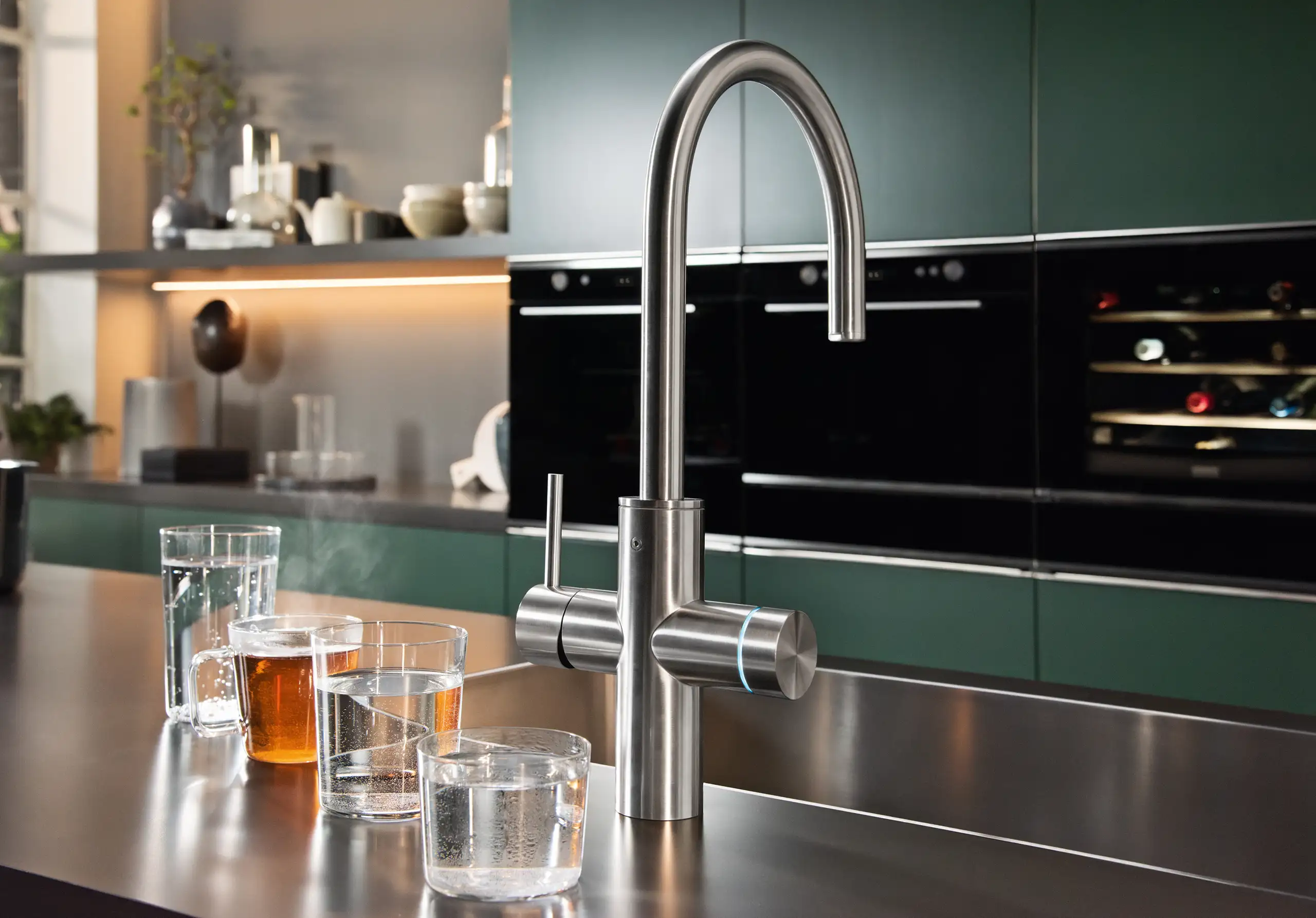 Eine moderne Küche mit grünlichen Schränken im Hintergrund. Im Vordergrund steht ein Edelstahl-Wasserhahn auf einer Arbeitsplatte. Daneben sind vier mit Flüssigkeiten gefüllte Gläser.