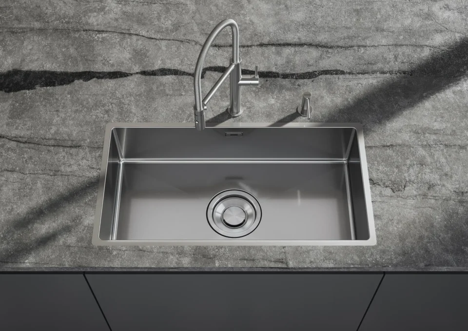 Ein rechteckiges, modernes Edelstahlspülbecken ist in eine graue Steinplatte eingelassen. Es hat einen hohen, gebogenen Wasserhahn und einen runden Abfluss in der Mitte.