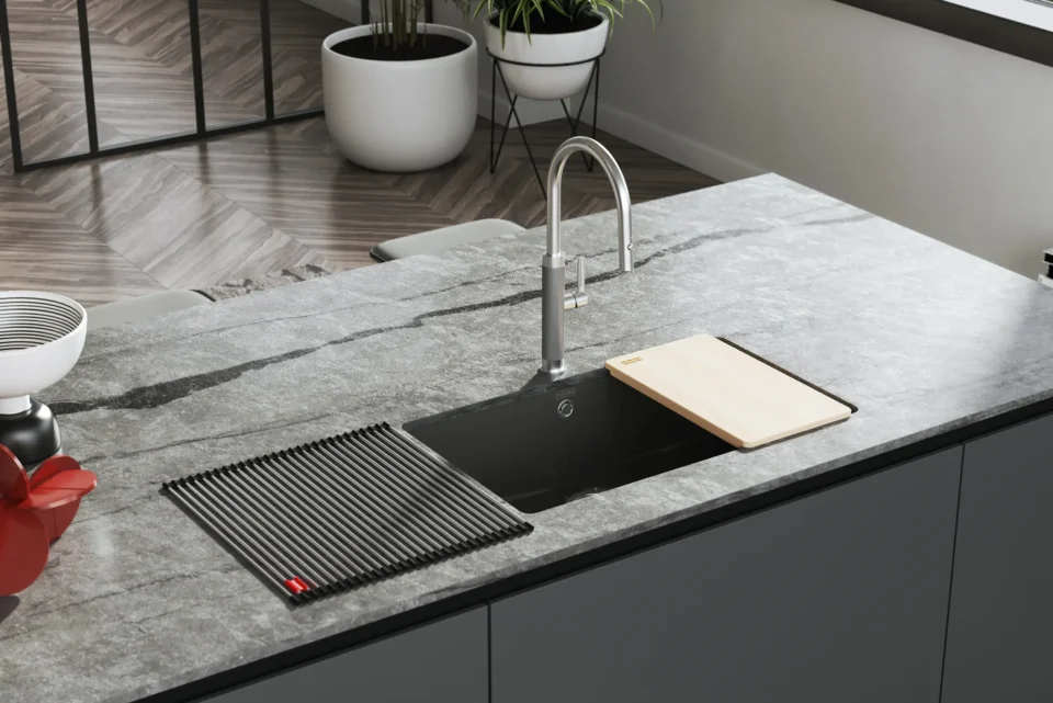 Eine moderne Küche mit einer grauen Marmorarbeitsplatte. Eine schwarze Spüle mit einem hohen Wasserhahn ist zu sehen, darauf ein Schneidebrett und ein Abtropfgestell. Im Hintergrund sind Pflanzen und Parkettboden.