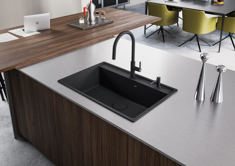 Eine moderne, offene Küche mit einer großen Kücheninsel. Die Insel hat eine schwarze Einbauspüle und einen gebogenen Wasserhahn. Im Hintergrund ist ein Holztisch mit grünen Stühlen und einem Laptop zu sehen.