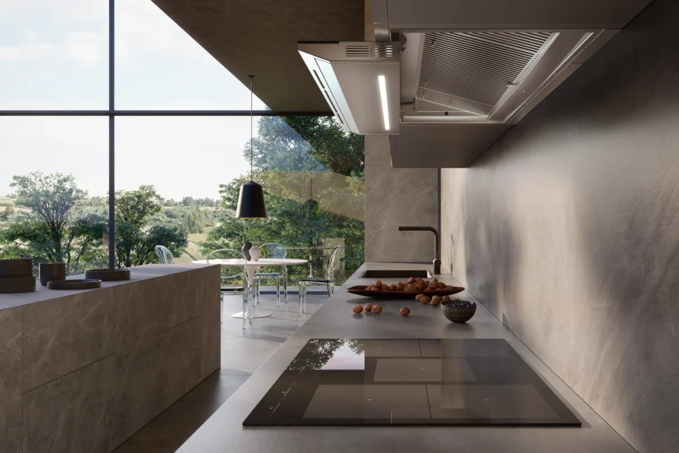 Moderne Küche mit minimalistischer Einrichtung, großen Fenstern und Blick auf einen grünen Garten. Helle Arbeitsplatten mit integriertem Kochfeld, einigen Schalen und einer schwarzen Hängelampe im Vordergrund.