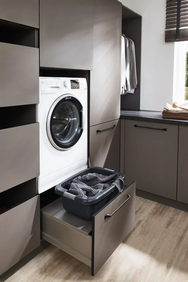 Moderner Hauswirtschaftsraum mit grauen Schränken, einer eingebauten Waschmaschine und einer offenen Schublade, in der ein Wäschekorb mit Kleidung steht, daneben ein Regal mit gefalteten Handtüchern. Der Boden ist aus Holz.