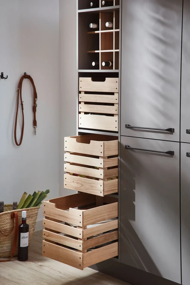 Ein Küchenschrank mit hölzernen Schubladen ist offen. Drei Schubladen sind sichtbar: zwei oben, eine unten. Im oberen Fach sind Flaschenhalterungen. Davor steht ein Korb mit Lebensmitteln und eine Flasche Balsamico.