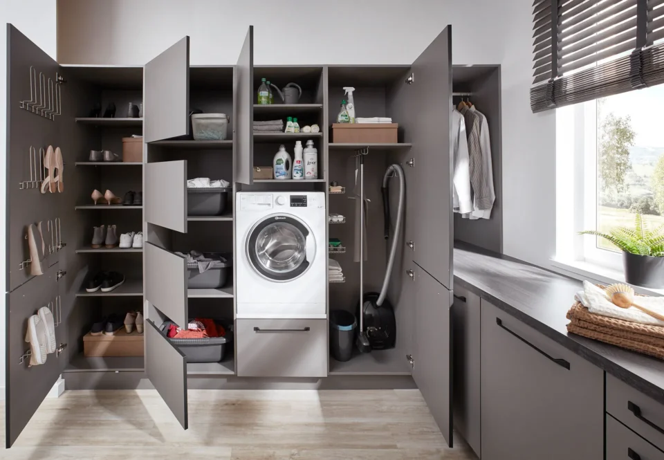 Eine aufgeräumte Waschküche mit offenen grauen Schränken. In einem Schrank befindet sich eine Waschmaschine, daneben Reinigungsmittel, Schuhe, ein Staubsauger und Haushaltsgegenstände. Tageslicht strömt durch ein Fenster.