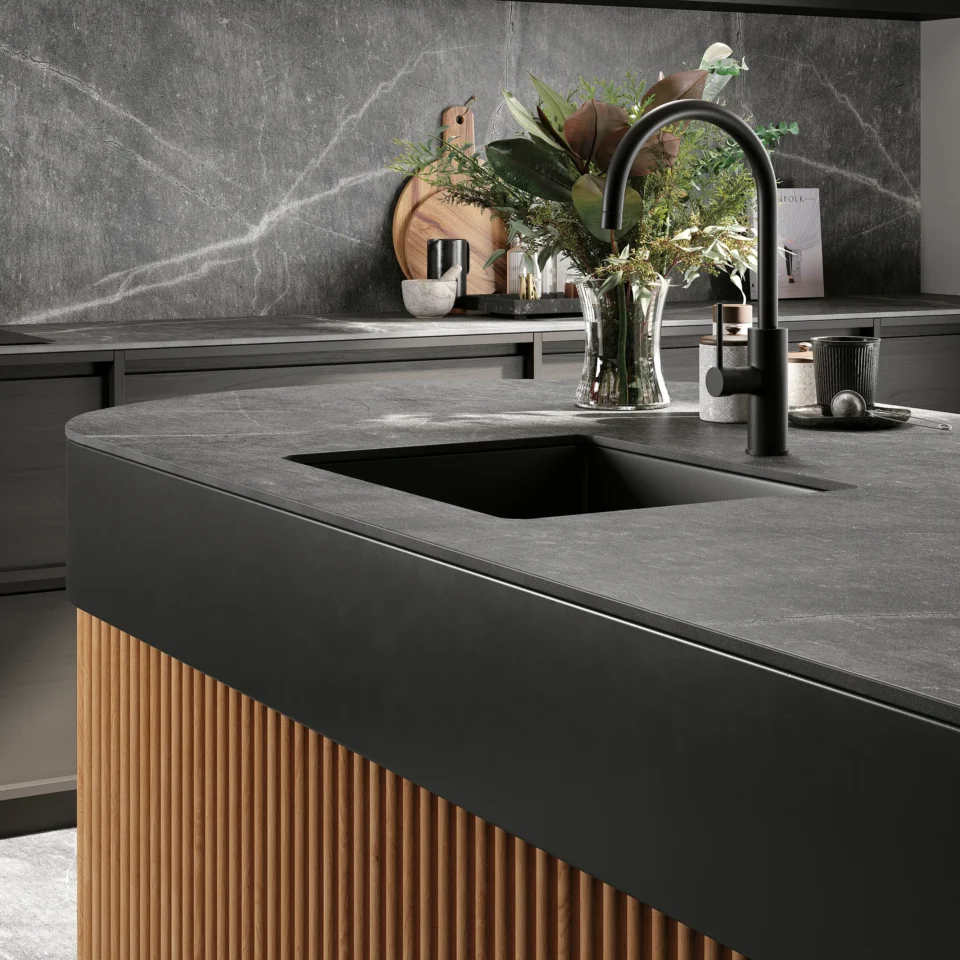 Moderne Küche mit dunkler Arbeitsplatte und Spüle. Neben der Spüle steht eine Vase mit grünen und roten Pflanzen. Die Schränke sind aus grauem Holz, die Rückwand aus grauem Stein mit weißen Aderungen.