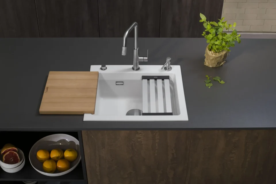 Eine schwarze Küchenarbeitsplatte mit einem weißen Spülbecken. Daneben ein Holzschneidebrett und ein Abtropfgitter. Eine silberne Armatur ist zu sehen. Oben rechts steht ein Topf mit einer Pflanze. Links unten sind Obst in einer Schale.