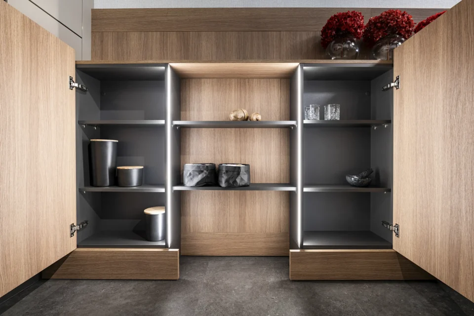 Ein offener Küchenschrank mit Holzoptik zeigt mehrere Regalböden. Darin befinden sich Vorratsdosen, Gläser, zwei schwarze Beutel und im oberen Fach der Mitte liegen zwei Knoblauchzehen. Oben rechts stehen rote Blumen.